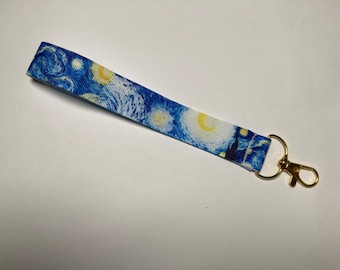 May include: A blue wrist lanyard featuring a print of Van Gogh's "Starry Night" painting. The lanyard has a gold-colored metal clasp for attaching keys or other items. The design includes swirling blue, yellow, and white elements.