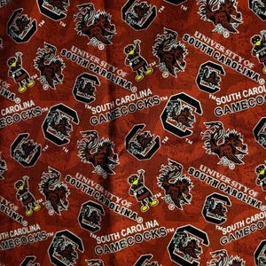 May include: Red fabric with a repeating pattern of the University of South Carolina Gamecocks logo, including the mascot, the wordmark, and the school name. The design is in black and white on a red background.