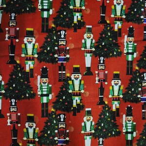 May include: Red fabric with a repeating pattern of Christmas nutcrackers and evergreen trees. The nutcrackers are in various colors, including green, red, and white, with black hats and gold accents. The trees are dark green.