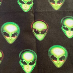 May include: Black fabric with a repeating pattern of green alien faces. The aliens have large, oval eyes and a smooth, green skin tone. Some alien faces have a red outline around the eyes. The fabric is likely used for apparel or home decor.