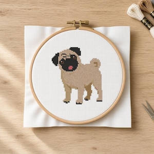 May include: A cross-stitch project featuring a tan pug with a black face and a curled tail, centered within a round wooden embroidery hoop on white fabric. Three skeins of embroidery floss in white, tan, and gray sit nearby with gold scissors.