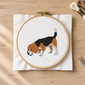 May include: A cross-stitch project in a round wooden embroidery hoop featuring a pixelated beagle dog with black, brown, and white fur. The design is stitched onto a square piece of white Aida fabric, showcasing the dog in a standing position.