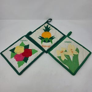 May include: Three square quilted pot holders with green fabric trim and hanging loops. Each white pot holder features embroidered tropical designs and the word Hawaii, including a pineapple, hibiscus flowers, and yellow lilies.