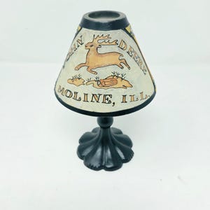 May include: A small table lamp with a black metal base and a conical shade. The shade features a tan deer illustration, the text JOHN DEERE, and the text MOLINE, ILL. The lamp has a black rim at the top and bottom of the shade.