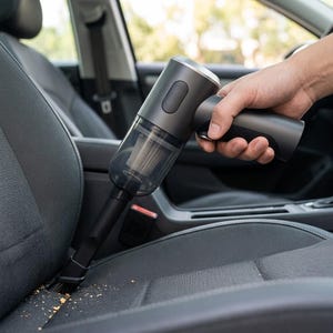 May include: A handheld cordless vacuum cleaner with a matte dark gray finish and a transparent dust canister. The device features a slim nozzle attachment currently cleaning debris from the textured fabric surface of a dark gray car seat.