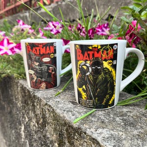 Pode incluir: Duas canecas de cerâmica branca com arte de capa de banda desenhada de The Batman. A caneca da frente mostra uma personagem com casaco verde e máscara com um símbolo de ponto de interrogação. A caneca de trás mostra uma figura escura.