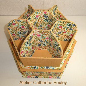 Peut inclure: Un organisateur de bureau hexagonal à compartiments multiples avec un motif floral de fleurs roses, bleues et de feuilles vertes sur fond blanc. L'extérieur est de couleur beige uni et le texte Atelier Catherine Bouley est inscrit en bas.