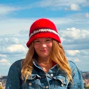 May include: A bright red knit bucket hat featuring a horizontal band with a white and dark brown striped pattern. The hat has a soft, fuzzy texture and a rounded crown with a short, downward-sloping brim that frames the head.