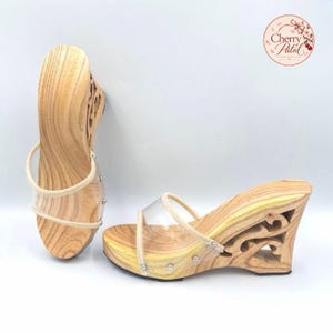 May include: A pair of light-coloured wooden wedge sandals featuring a natural wood grain pattern, clear plastic straps, and a decorative cutout design on the side of the wedge heel. The sandals are accented with small silver-toned metal studs.