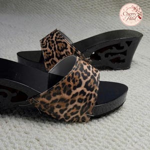 Könnte beinhalten: Ein Paar Schlupf-Keilsandalen mit einem Obermaterial aus Stoff mit Leopardenmuster und einer schwarzen, glänzenden Basis. Die Keilabsätze haben ein dekoratives Ausschnittdesign. Ein kreisförmiges Logo oben rechts zeigt den Text Cherry Patal.