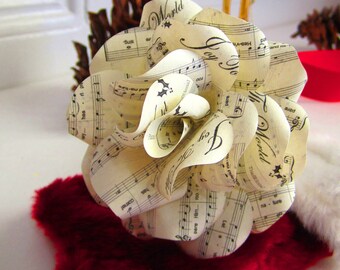 May include: A white paper flower made from sheet music. The flower is shaped like a rose and has a delicate, vintage look. The music sheets have the words "Joy to the World" and "Let it Snow" printed on them.