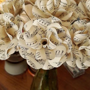 May include: A bouquet of paper flowers crafted from vintage sheet music. The flowers are cream-colored with black musical notes, arranged in a green glass vase. The petals are carefully curled to resemble roses.