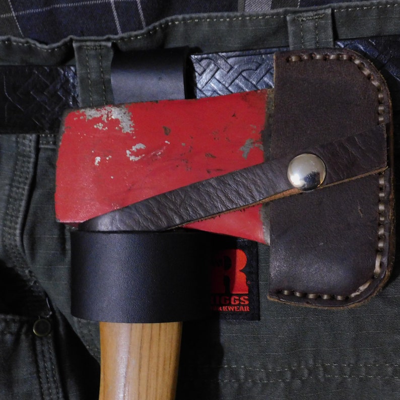 May include: A red axe head with a worn appearance is secured in a brown leather sheath. A black leather belt and a black leather loop hold the axe in place. The axe handle is made of wood. The axe is attached to a pair of green pants.
