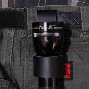 May include: A black Mag-Lite flashlight secured in a black leather belt loop, attached to a green work pants. The flashlight is partially inserted into a black leather holder. The belt is black, with a textured pattern.