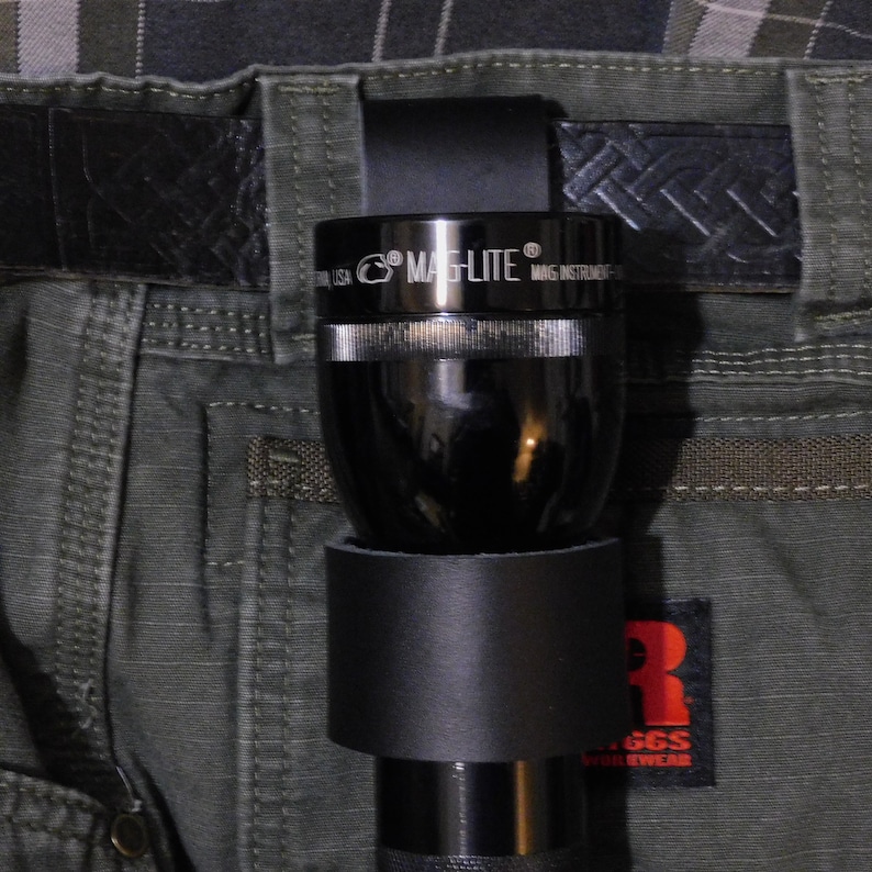 May include: A black Mag-Lite flashlight secured in a black leather belt loop, attached to a green work pants. The flashlight is partially inserted into a black leather holder. The belt is black, with a textured pattern.