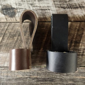 May include: Two leather cup holders, one brown and one black, sit side-by-side. Each has a loop handle and a cylindrical base. The brown holder has a lighter brown loop, while the black holder has a black loop. The items are on a wooden surface.
