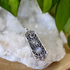 May include: A silver pendant necklace with a moonstone and a detailed design of a crescent moon, stars, and a sun. The pendant is hanging on a white rock.