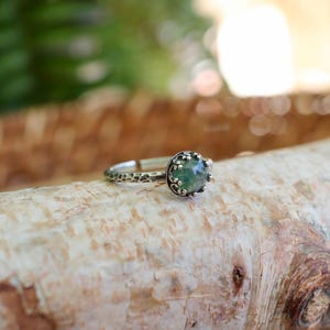 Puede incluir: Un anillo de plata con una piedra preciosa redonda de color verde moteado, engastada en un bisel de metal texturizado con cuentas. La banda del anillo tiene un acabado martillado e irregular, lo que le da un aspecto t&aacute;ctil a la estructura.