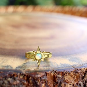 May include: A gold-toned ring featuring a central starburst design with a round, iridescent opal stone. The band is decorated with small engraved star motifs on either side of the main starburst setting, creating a celestial-inspired appearance.
