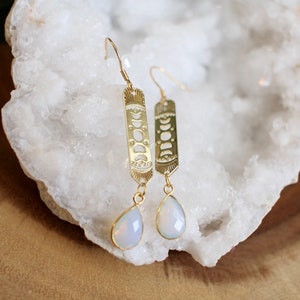 May include: Gold-colored earrings with a celestial design. Each earring features a rectangular pendant with a moon phase pattern and a teardrop-shaped, opalescent stone. The earrings are suspended from a gold-colored hook.