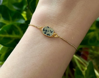 May include: A gold chain bracelet with a small, oval-shaped dalmatian jasper stone pendant. The stone is a light green color with black spots.