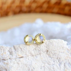 May include: A pair of small, round gold earrings with a textured border and a light blue center. The earrings are displayed on a white, textured surface, with a blurred green and brown background.