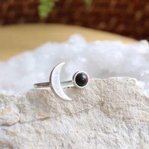 May include: A silver ring featuring a crescent moon and a small, round, dark gemstone. The ring is set on a white, textured surface, with green foliage in the background. The ring's design is simple and elegant.