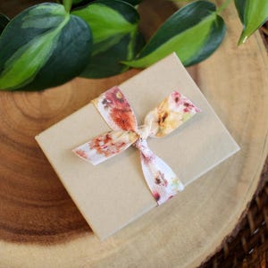 May include: A small, tan gift box tied with a floral ribbon. The ribbon features a pattern of red, orange, and yellow flowers. The box is resting on a wooden surface, with green leaves in the background.