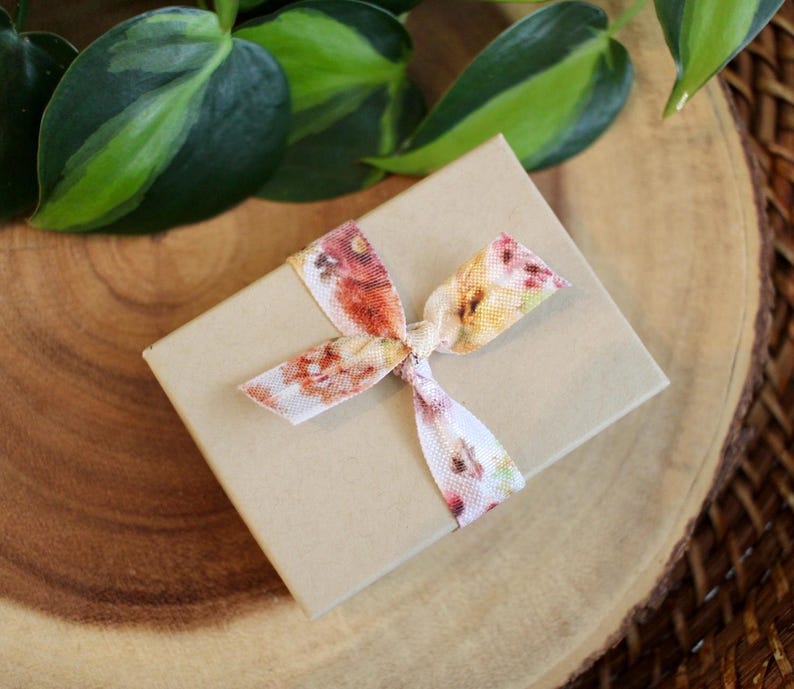 May include: A small, tan gift box tied with a floral ribbon. The ribbon features a pattern of red, orange, and yellow flowers. The box is resting on a wooden surface, with green leaves in the background.