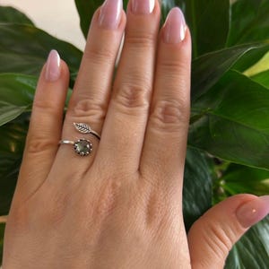 May include: A silver ring with a leaf design and a round, gray gemstone. The ring is worn on a hand with light pink nail polish and is set against a background of green leaves.
