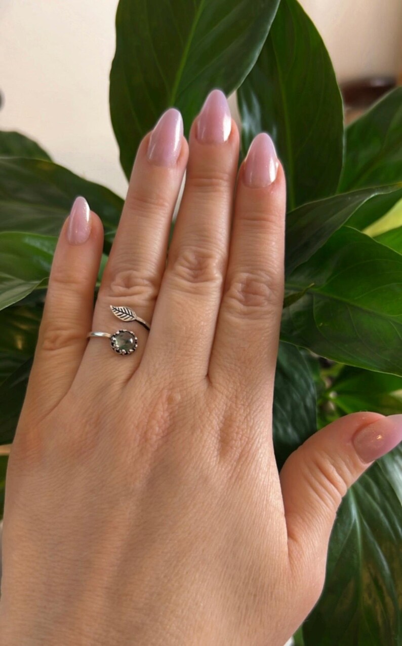 May include: A silver ring with a leaf design and a round, gray gemstone. The ring is worn on a hand with light pink nail polish and is set against a background of green leaves.