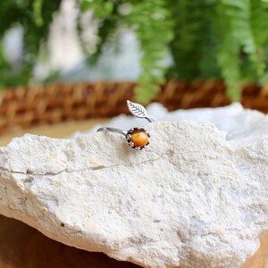 May include: A silver-toned ring with an adjustable band, featuring a small, round, orange gemstone surrounded by tiny clear stones. A delicate leaf design accents the band. The ring is displayed on a white rock against a blurred green and brown background.