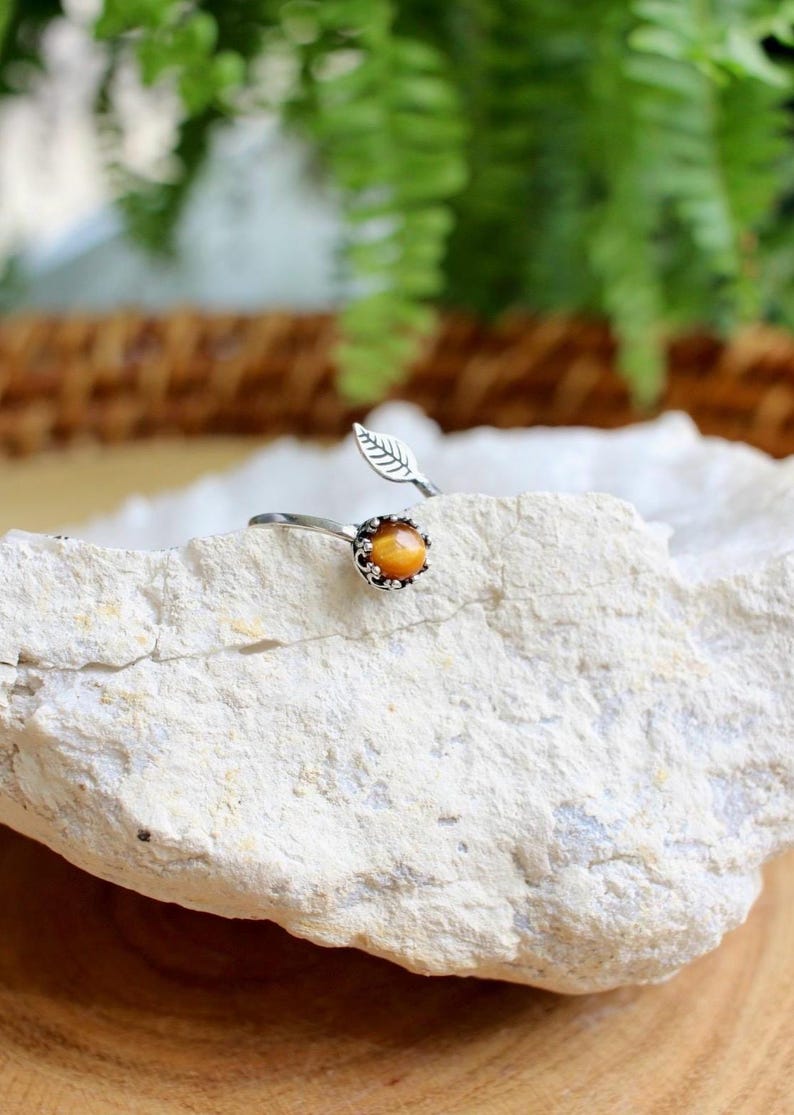 May include: A silver-toned ring with a leaf design and a round, orange gemstone. The ring is set on a white rock, with green foliage in the background. The gemstone is approximately 0.5 inches in diameter.