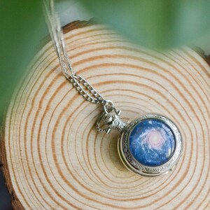 Puede incluir: Un reloj de bolsillo plateado con una cadena cuelga de una superficie de madera. La esfera del reloj presenta un dise&ntilde;o de galaxia azul y rosa.