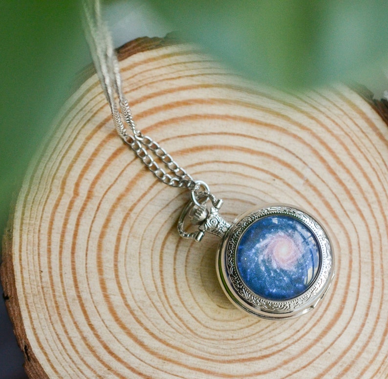 Puede incluir: Un reloj de bolsillo plateado con una cadena cuelga de una superficie de madera. La esfera del reloj presenta un dise&ntilde;o de galaxia azul y rosa.