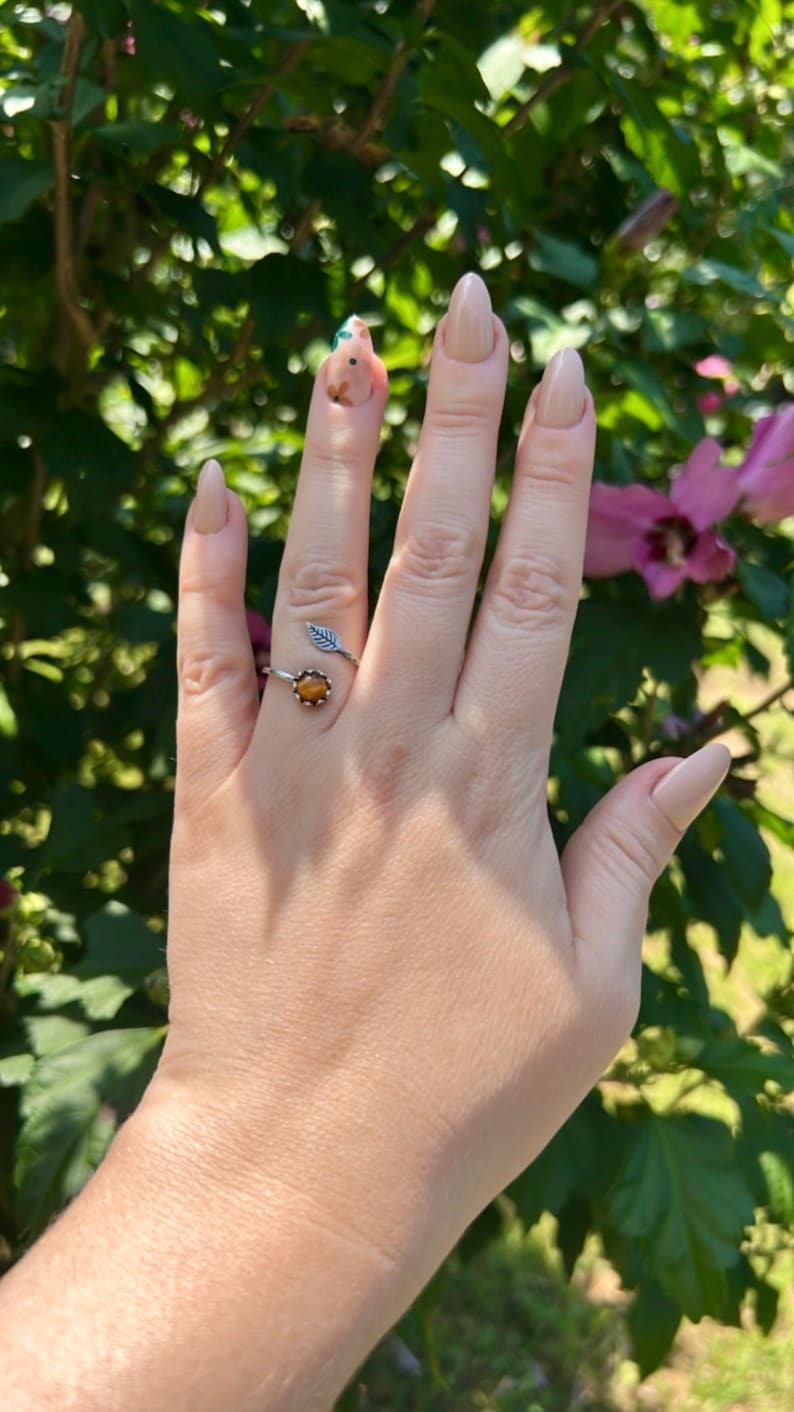May include: A silver ring with a leaf design and a round, amber-colored stone. The ring is on a hand with long, polished nails. The background is blurred green foliage and pink flowers.
