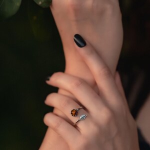 May include: Two silver rings are displayed on a hand. One ring features a round, orange gemstone. The other ring has a dark blue gemstone. The rings are set against a dark green background.