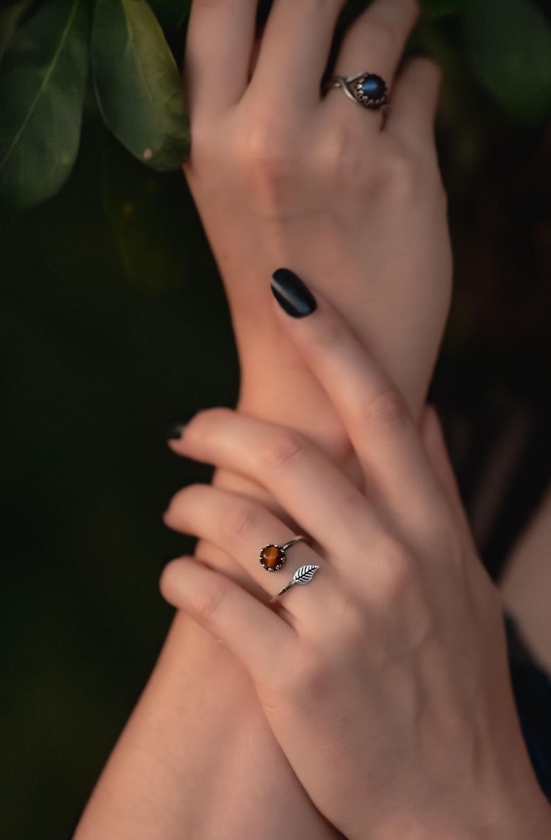 May include: Two silver rings are displayed on a hand. One ring features a round, orange gemstone. The other ring has a dark blue gemstone. The rings are set against a dark green background.