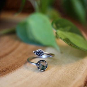 May include: A silver-toned ring with a leaf design. The ring features a round, bezel-set stone with green and brown inclusions. The band is adjustable, with a leaf detail on one end.