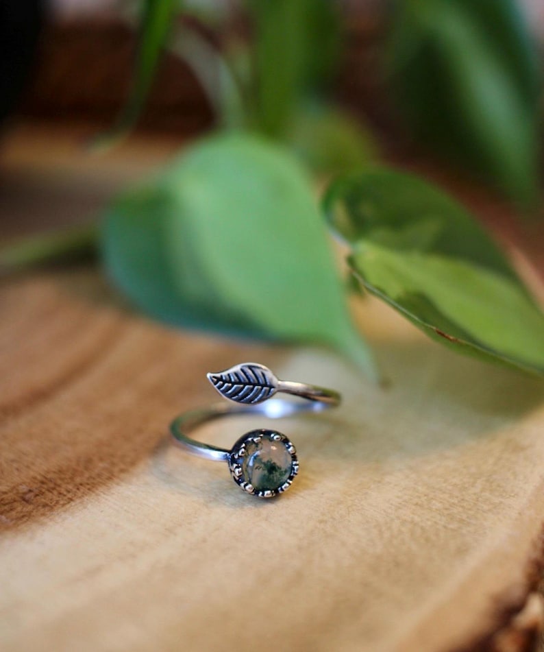 May include: A silver-toned ring with a leaf design. The ring features a round, bezel-set stone with green and brown inclusions. The band is adjustable, with a leaf detail on one end.
