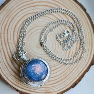 Puede incluir: Un reloj de bolsillo plateado con una cadena. La esfera del reloj presenta un dise&ntilde;o de galaxia azul y rosa.