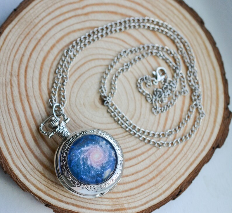 Puede incluir: Un reloj de bolsillo plateado con una cadena. La esfera del reloj presenta un dise&ntilde;o de galaxia azul y rosa.