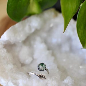 May include: A silver ring with a round, green stone and a leaf design. The ring is set on a white, textured surface, with green leaves in the background. The ring's design is nature-inspired.