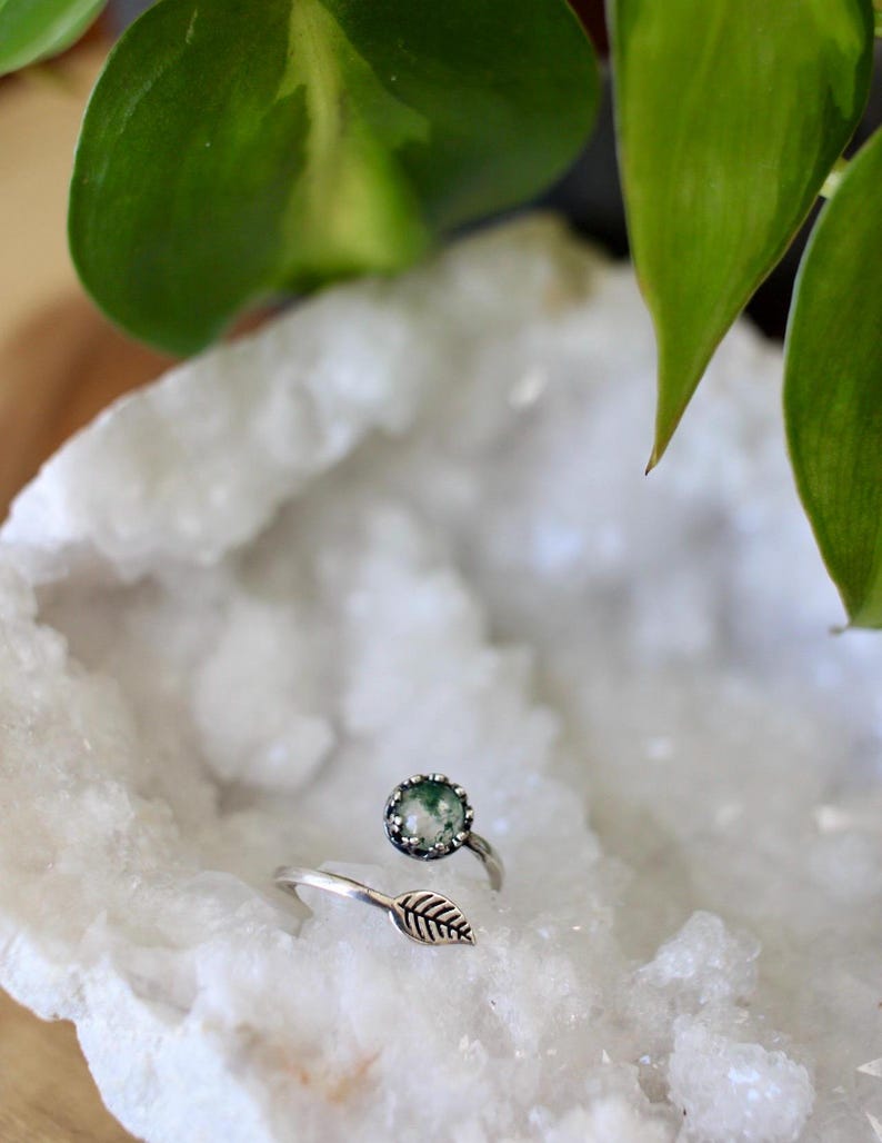 May include: A silver ring with a round, green stone and a leaf design. The ring is set on a white, textured surface, with green leaves in the background. The ring's design is nature-inspired.