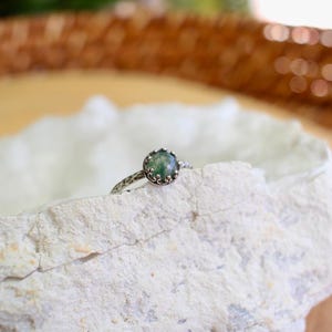 Puede incluir: Un anillo de plata con una piedra preciosa redonda de color verde musgo en un engaste de garras en forma de corona. La banda del anillo tiene un acabado martillado y texturizado. El anillo est&aacute; colocado sobre el borde de una roca blanca.