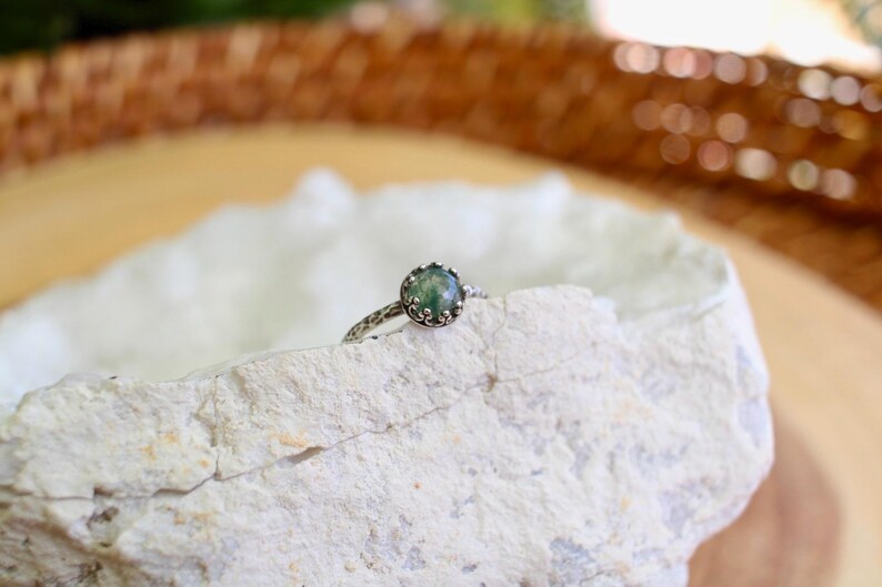 Puede incluir: Un anillo de plata con una piedra preciosa redonda de color verde musgo en un engaste de garras en forma de corona. La banda del anillo tiene un acabado martillado y texturizado. El anillo est&aacute; colocado sobre el borde de una roca blanca.