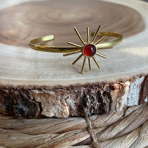 May include: A gold-toned cuff bracelet featuring a sunburst design with a central, round, orange-red stone. The sunburst has multiple rays extending outward from the center. The bracelet is displayed on a wooden surface.