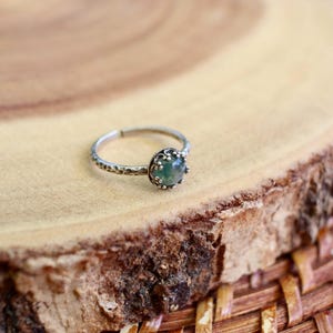 Puede incluir: Un anillo de tono plateado con una peque&ntilde;a piedra redonda de color verde transl&uacute;cido engastada en un bisel met&aacute;lico decorativo y festoneado. La banda del anillo tiene un acabado martillado texturizado y un dise&ntilde;o abierto ajustable.