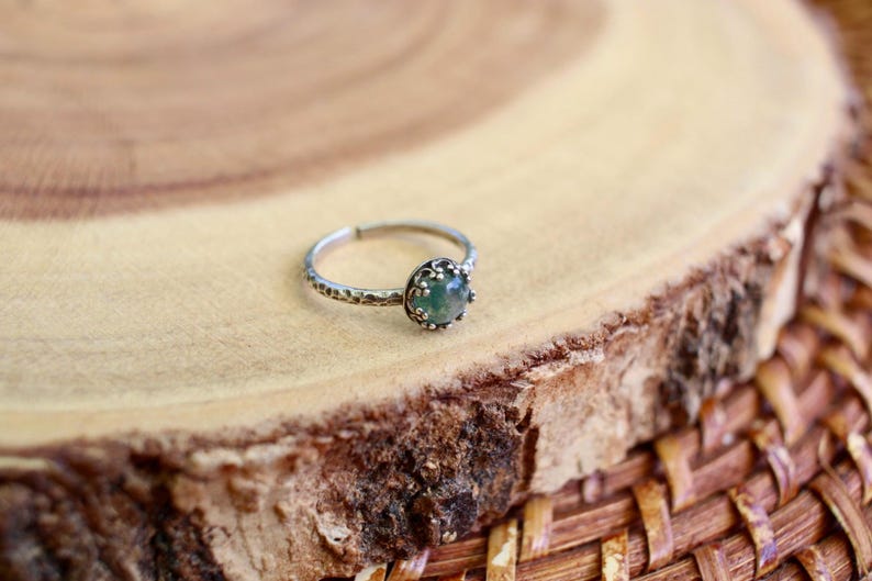 Puede incluir: Un anillo de tono plateado con una peque&ntilde;a piedra redonda de color verde transl&uacute;cido engastada en un bisel met&aacute;lico decorativo y festoneado. La banda del anillo tiene un acabado martillado texturizado y un dise&ntilde;o abierto ajustable.