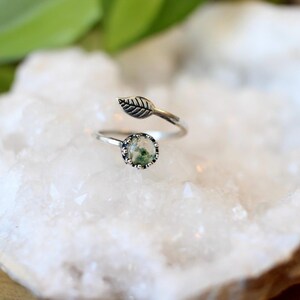 May include: A silver-toned ring with a leaf design and a round, green stone. The ring features an adjustable band and a small leaf detail. The stone has a unique, mossy appearance. The ring is set against a white, textured background.